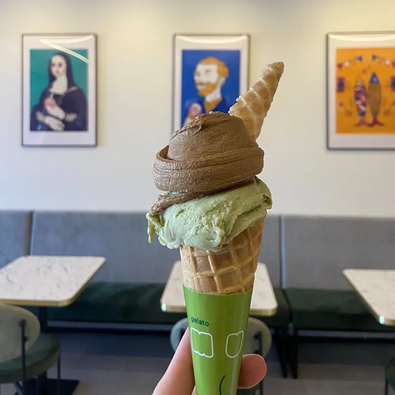 Uma mão segura um cone de gelato com duas bolas, embrulhado em papel verde da Musa. Atrás, a Mona Lisa e Van Gogh em quadros pop art.