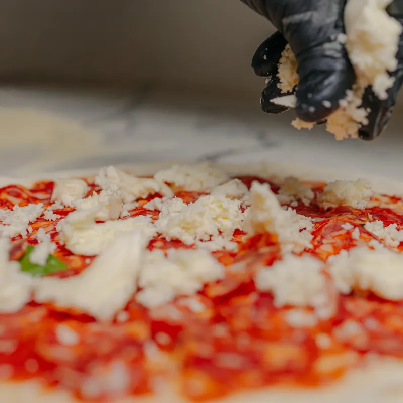 Mão de luva preta a espalhar mozzarella fior di latte sobre o tomate de uma pizza.