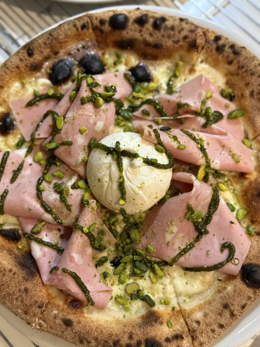 Pizza Musa vista de perto, com mortadela, uma burrata inteira ao centro, pesto e pistácio, e a borda tostada.