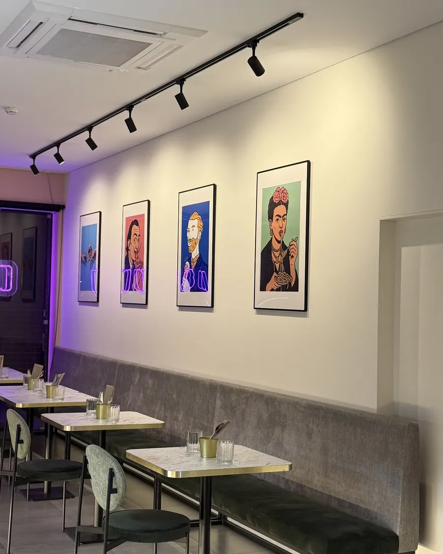 A sala comprida da Musa, com banco de veludo cinzento, mesas de mármore, cadeiras verde-sálvia e quadros pop art de pintores a comer.