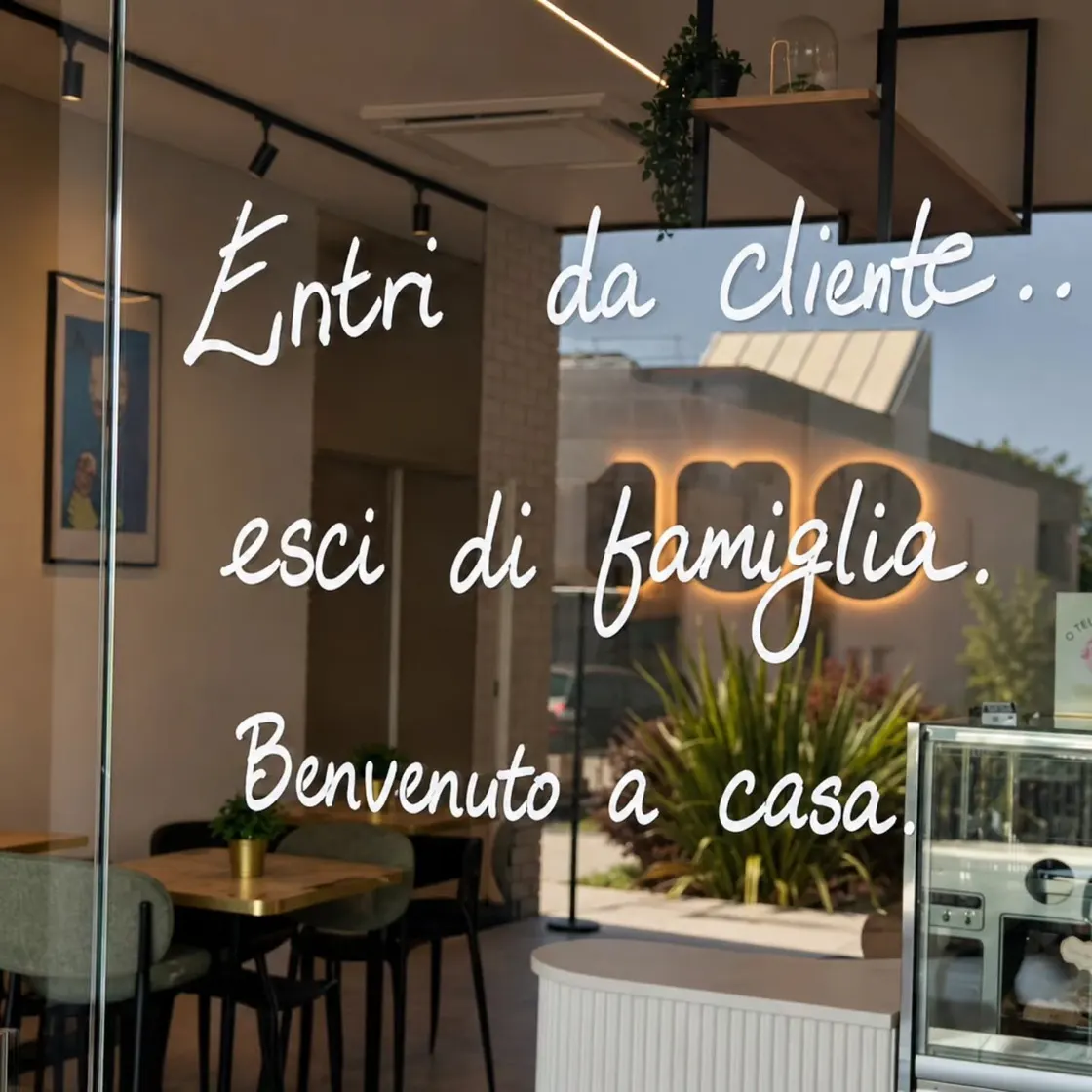 O vidro da Musa, escrito à mão: “Entri da cliente.. esci di famiglia. Benvenuto a casa.” Atrás, a sala.